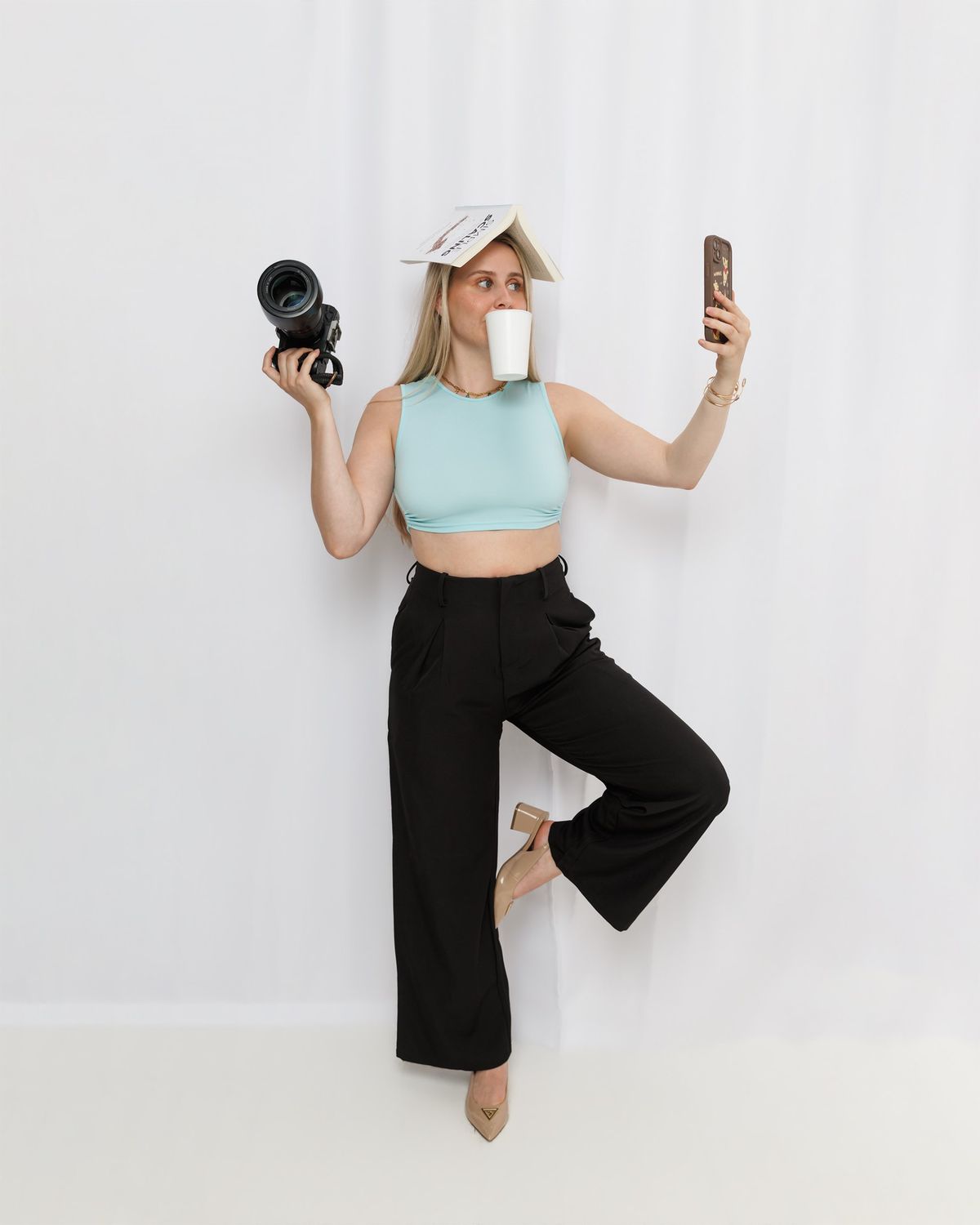 Victoria balancing a book on her head, a coffee cup in her mouth, a camera in one hand and a phone in the other